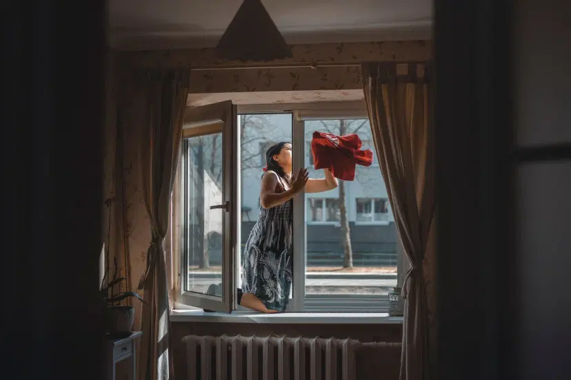 Woman standing on windowsill from inside, cleaning exterior window glass with red cloth. Beige curtains frame the open window, with bare trees and buildings visible outside on sunny day.