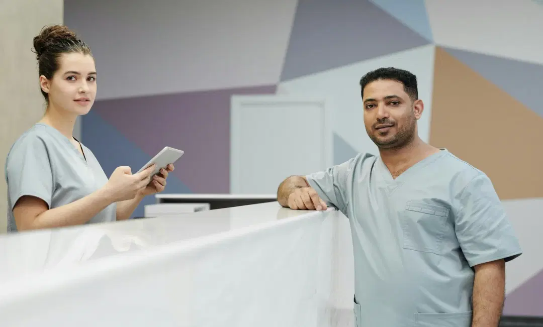 Female dental receptionist in light blue scrubs holding a tablet at a white reception desk, interacting with a male patient in matching scrubs in a modern dental office with geometric wall design.